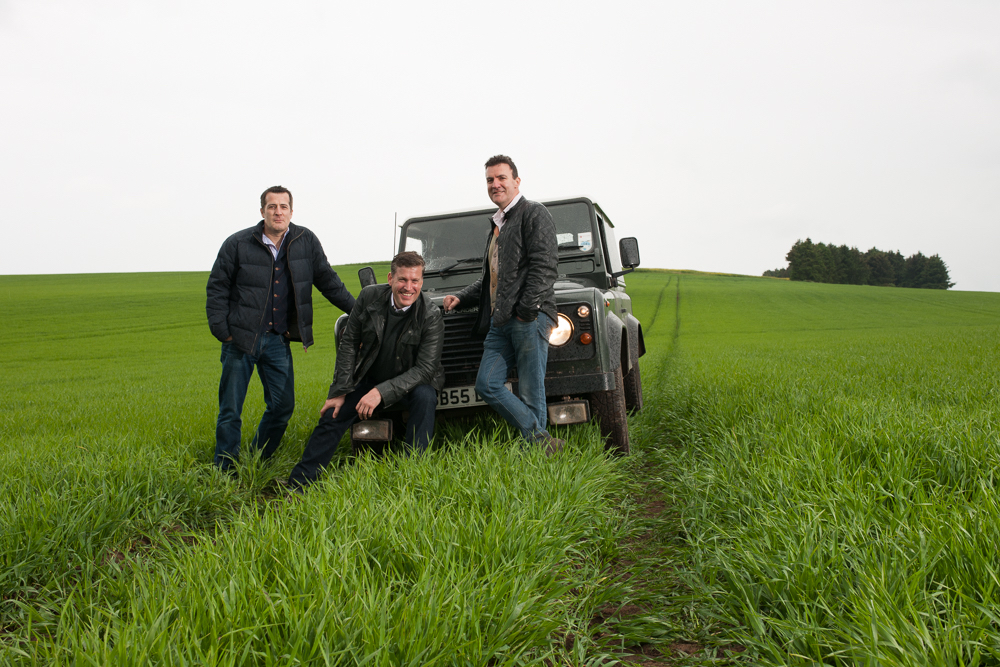 The owners of Arbikie Highland Estate, brothers Iain, David and John Stirling standing by a 4x4 vehicle in a field of grass.
