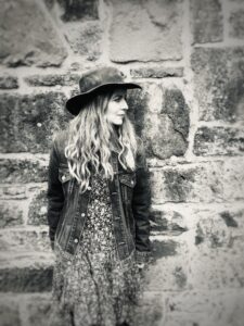 Black and white photo of artist Nicola Evans stood against a brick wall - with long wavy hair, dress, denim jacket and a black hat.