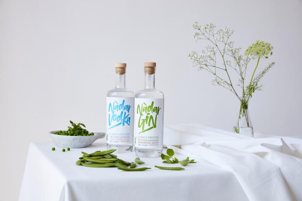 A bottle of potato-based vodka and a bottle of pea-based gin with the label Nàdar, on a table with a white tablecloth, bowl of peas and a vase of flowers. The bottles were produced by the Arbikie Highland Estate. 