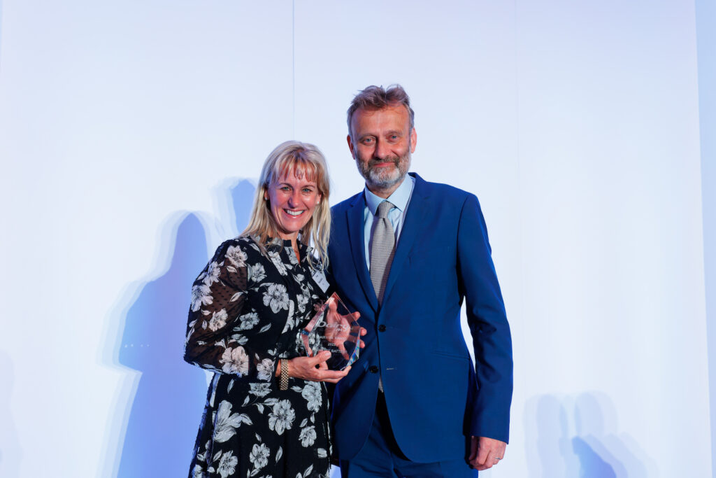 Minette Batters, president of the NFU, holding her Dairy UK award, standing against a white background with comedian and presenter Hugh Dennis
