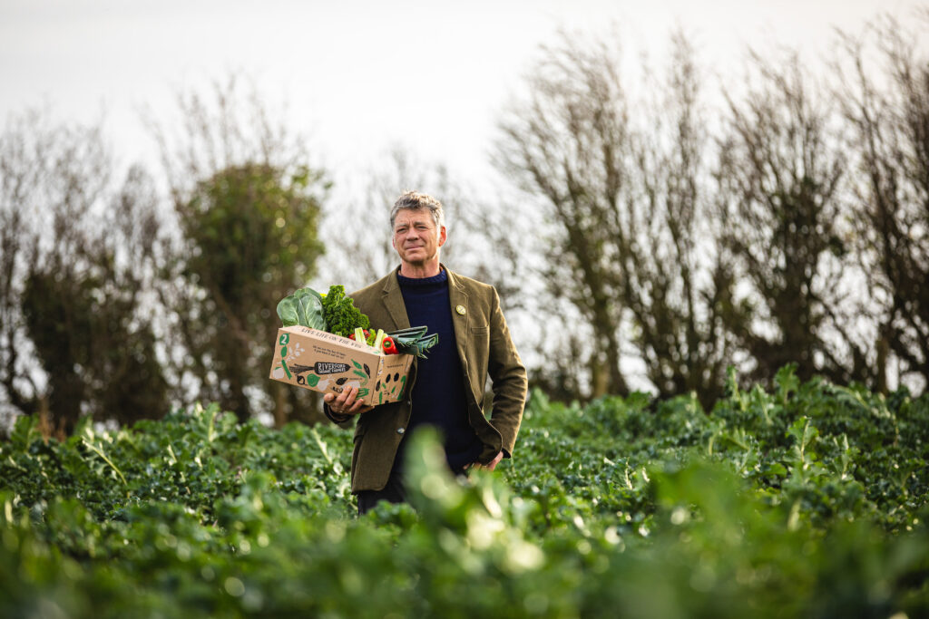 Guy Singh-Watson, founder of Riverford Organic, holding a box of fresh vegetables, sanding in a field.