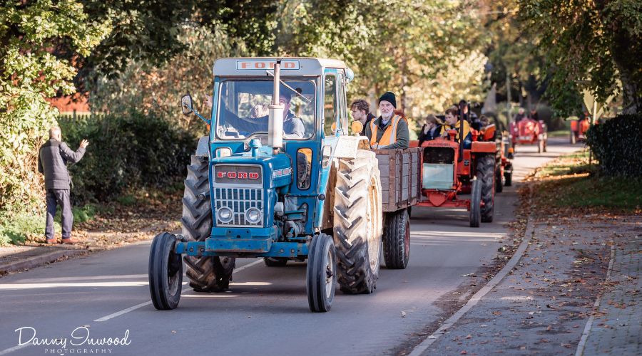 blue vintage Ford tractor followed by a line of tractors driving down a rural road lined with trees 